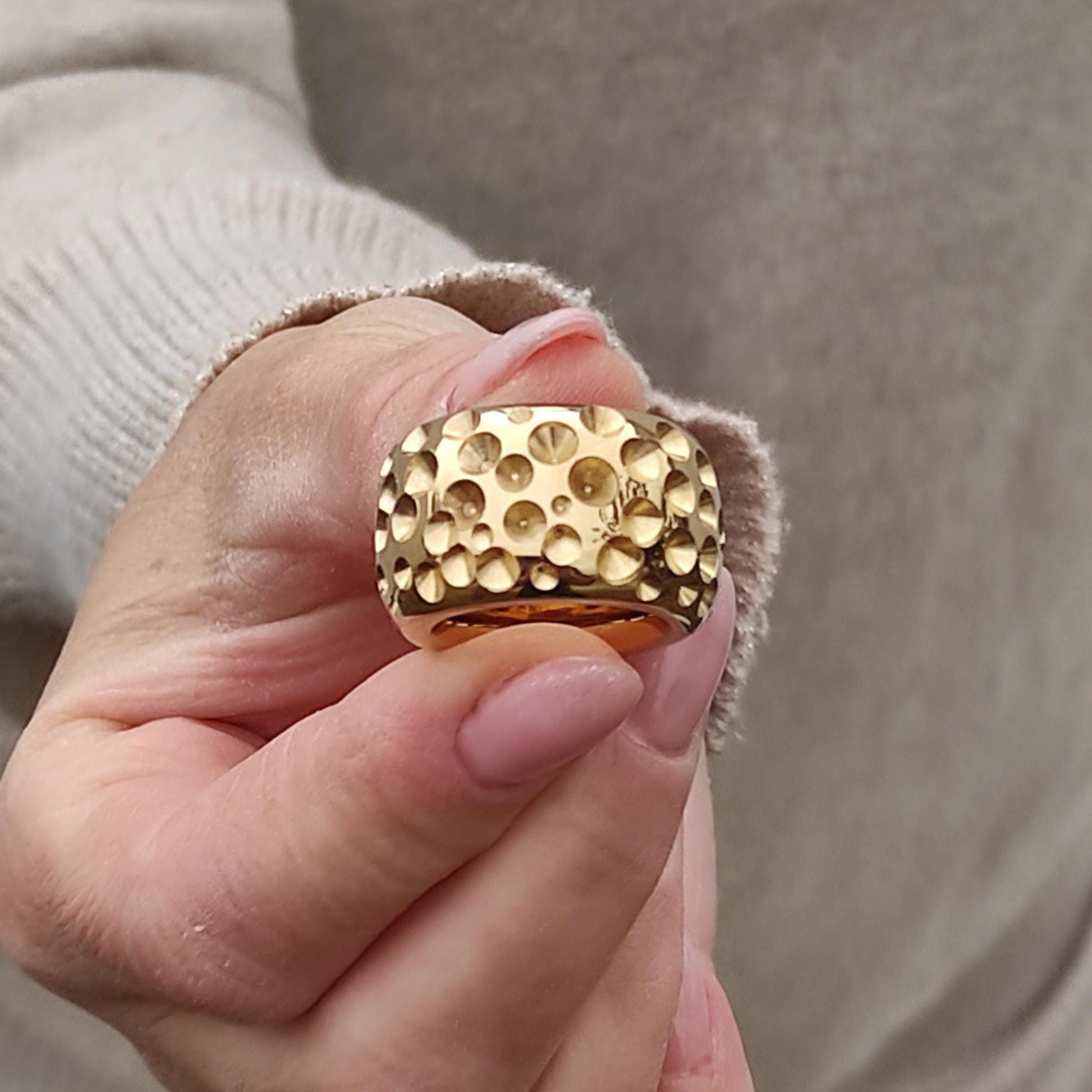 May include: Close-up of a gold ring with a textured surface. The ring has a wide band and a pattern of small, raised circles. The gold ring is a statement piece of jewelry.