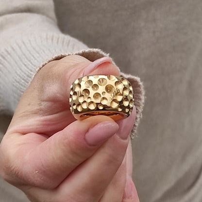 May include: Close-up of a gold ring with a textured surface. The ring has a wide band and a pattern of small, raised circles. The gold ring is a statement piece of jewelry.