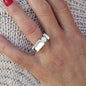 May include: A silver ring with a large, round, clear stone set in a square bezel. The ring has a thick band with a rectangular shape.