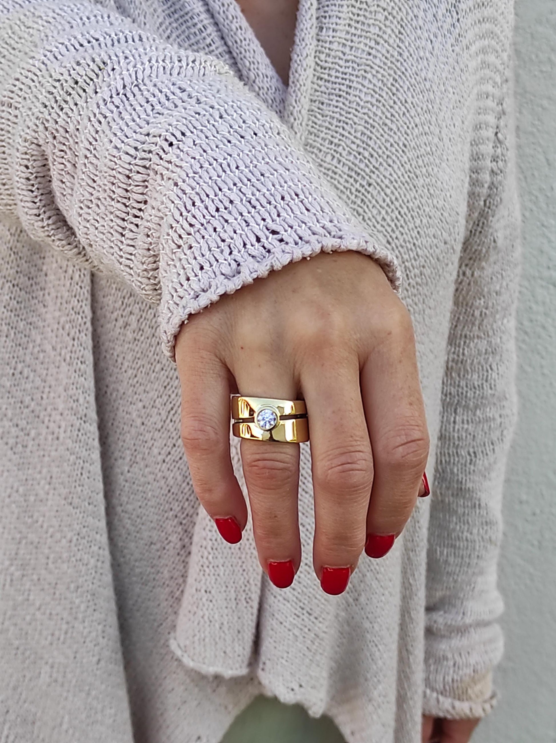 May include: A gold ring with a single, round, clear stone set in the center. The ring is made of two bands, one wider than the other, and is worn on the ring finger of a hand.