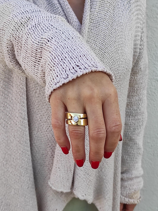 May include: A gold ring with a single, round, clear stone set in the center. The ring is made of two bands, one wider than the other, and is worn on the ring finger of a hand.