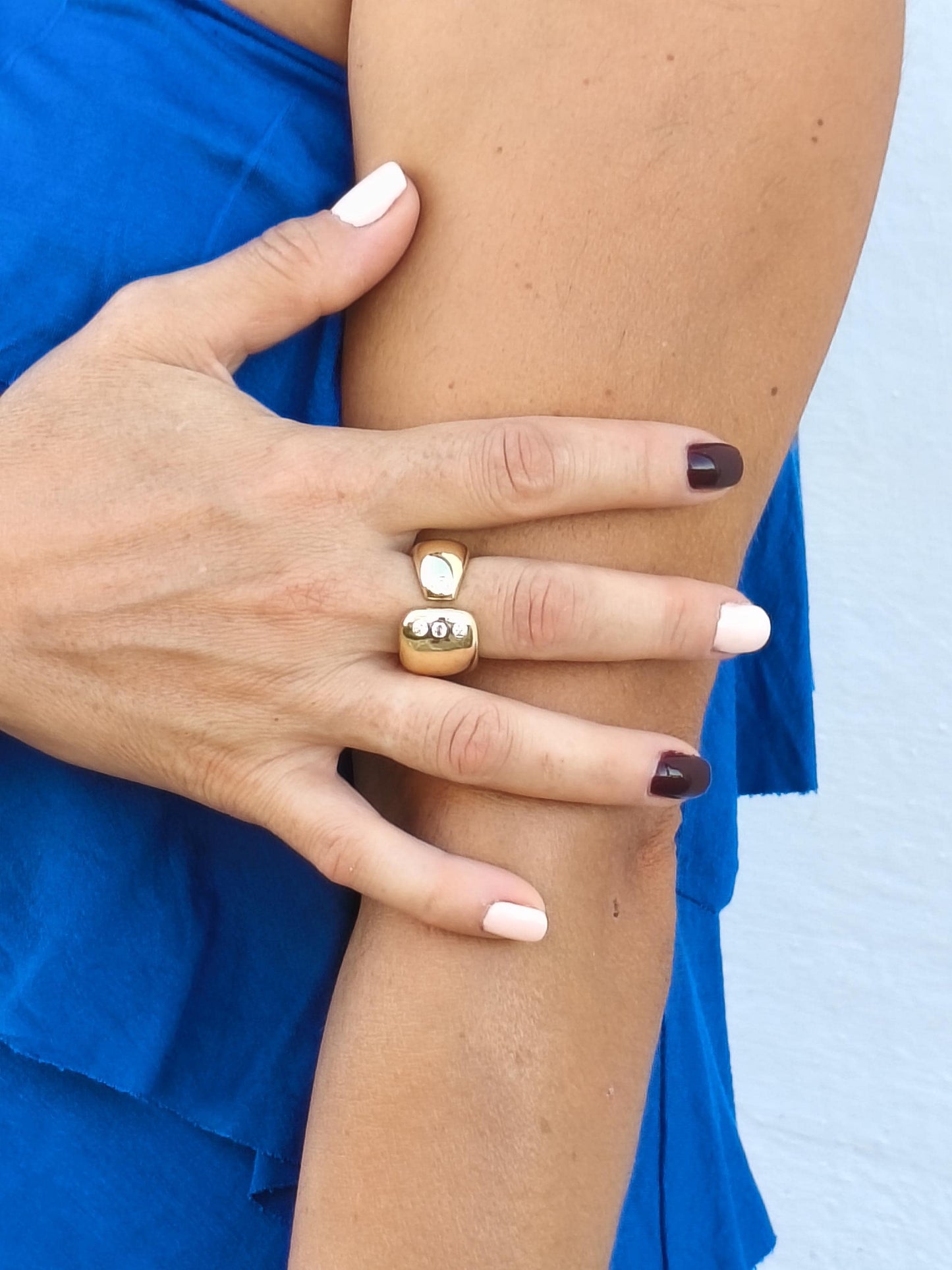 May include: A gold ring with a rectangular top and a smaller, rounded base. The top of the ring features three small, clear stones. The ring is worn on a finger with a manicure featuring white, light pink, and dark brown nail polish.