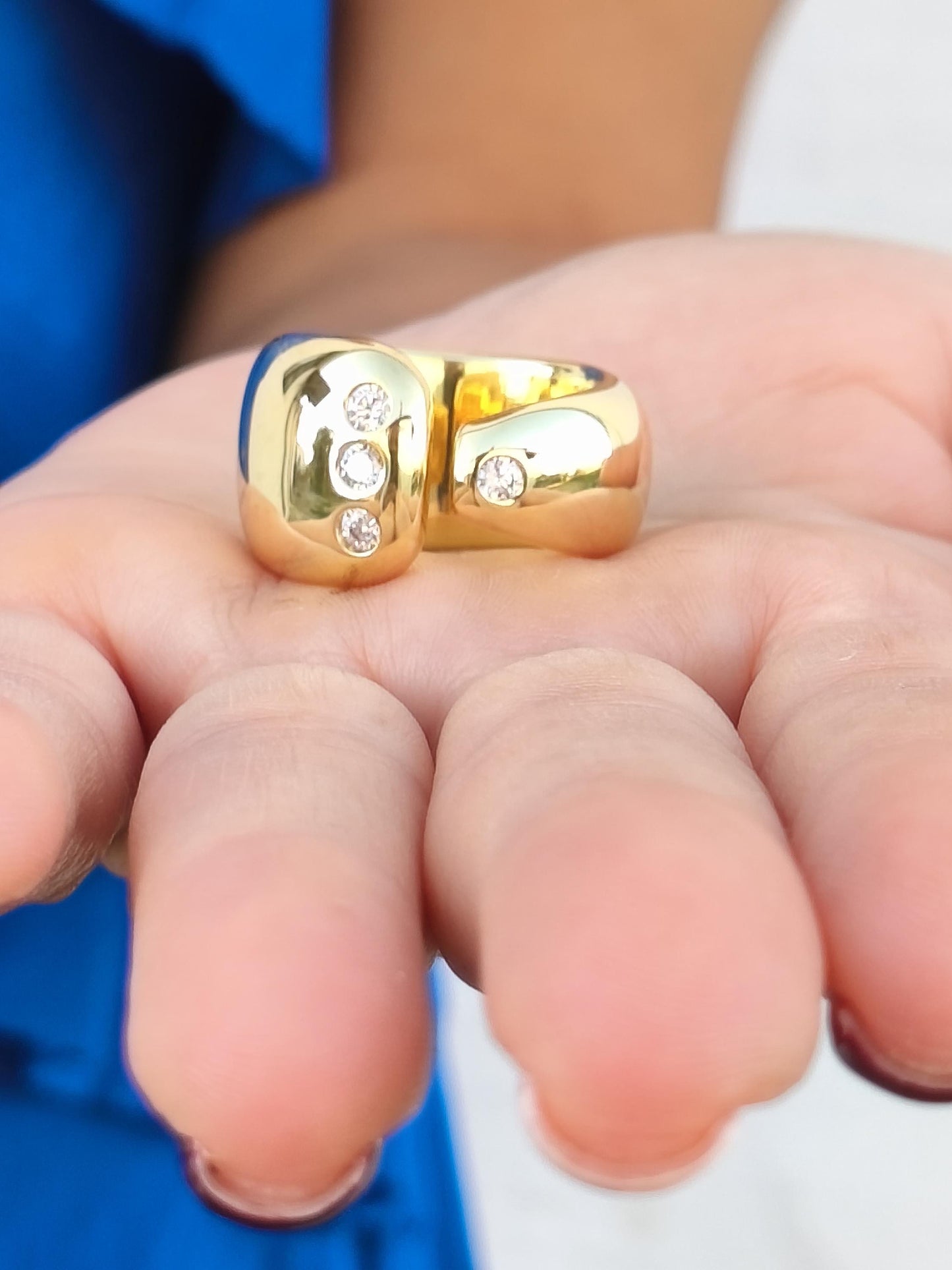 May include: A gold-colored ring with three small diamonds on one side and one on the other. The ring has a unique, curved design and is displayed on a person's open palm.