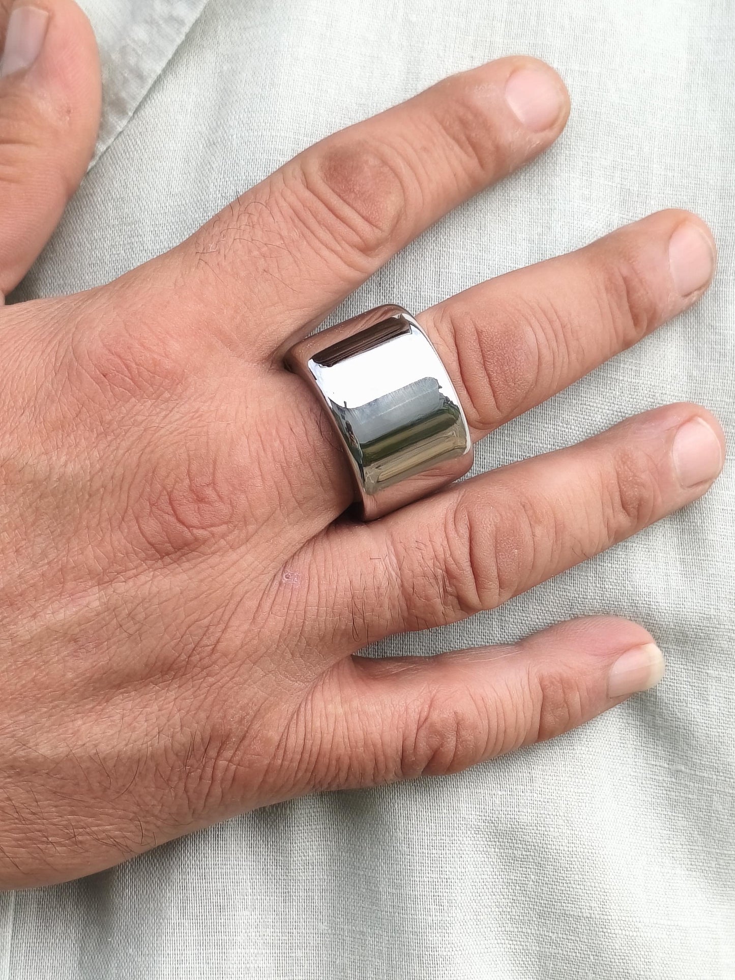 May include: A wide, polished silver ring with a rectangular design. The ring is worn on a finger, reflecting light. The jewelry piece has a smooth, shiny surface. This ring is a bold statement piece.