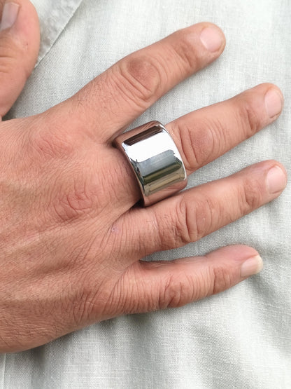 May include: A wide, polished silver ring with a rectangular design. The ring is worn on a finger, reflecting light. The jewelry piece has a smooth, shiny surface. This ring is a bold statement piece.