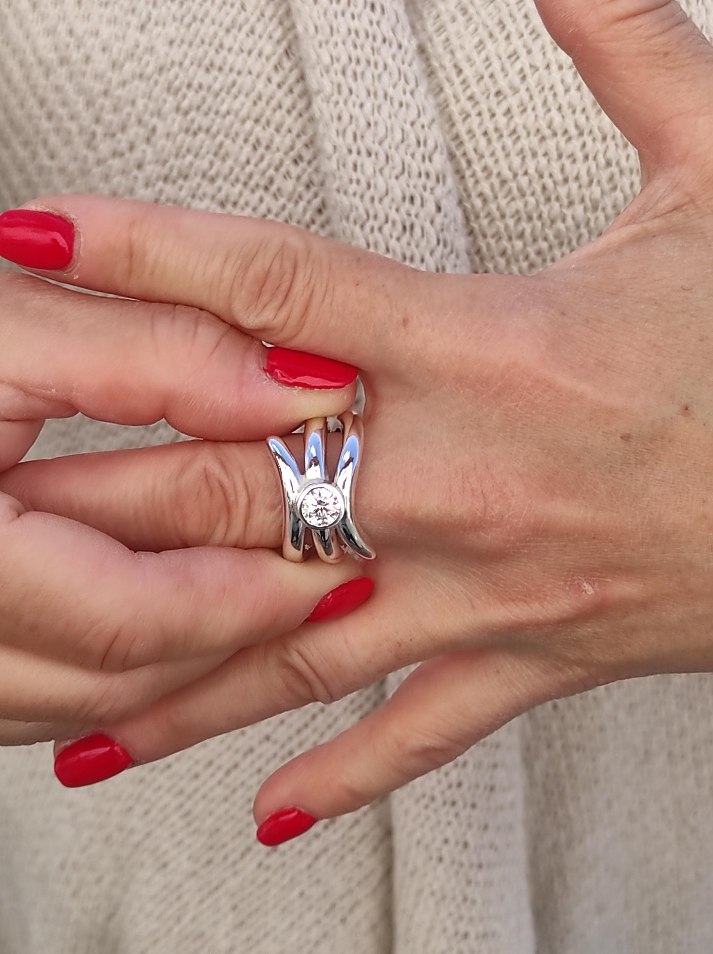 May include: A silver ring with a unique design featuring a central, clear gemstone. The ring has a modern, sculptural shape with curved, prong-like elements surrounding the stone. The ring is shown on a hand with red nail polish.