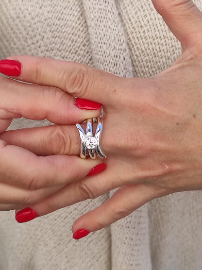 May include: A silver ring with a unique design featuring a central, clear gemstone. The ring has a modern, sculptural shape with curved, prong-like elements surrounding the stone. The ring is shown on a hand with red nail polish.
