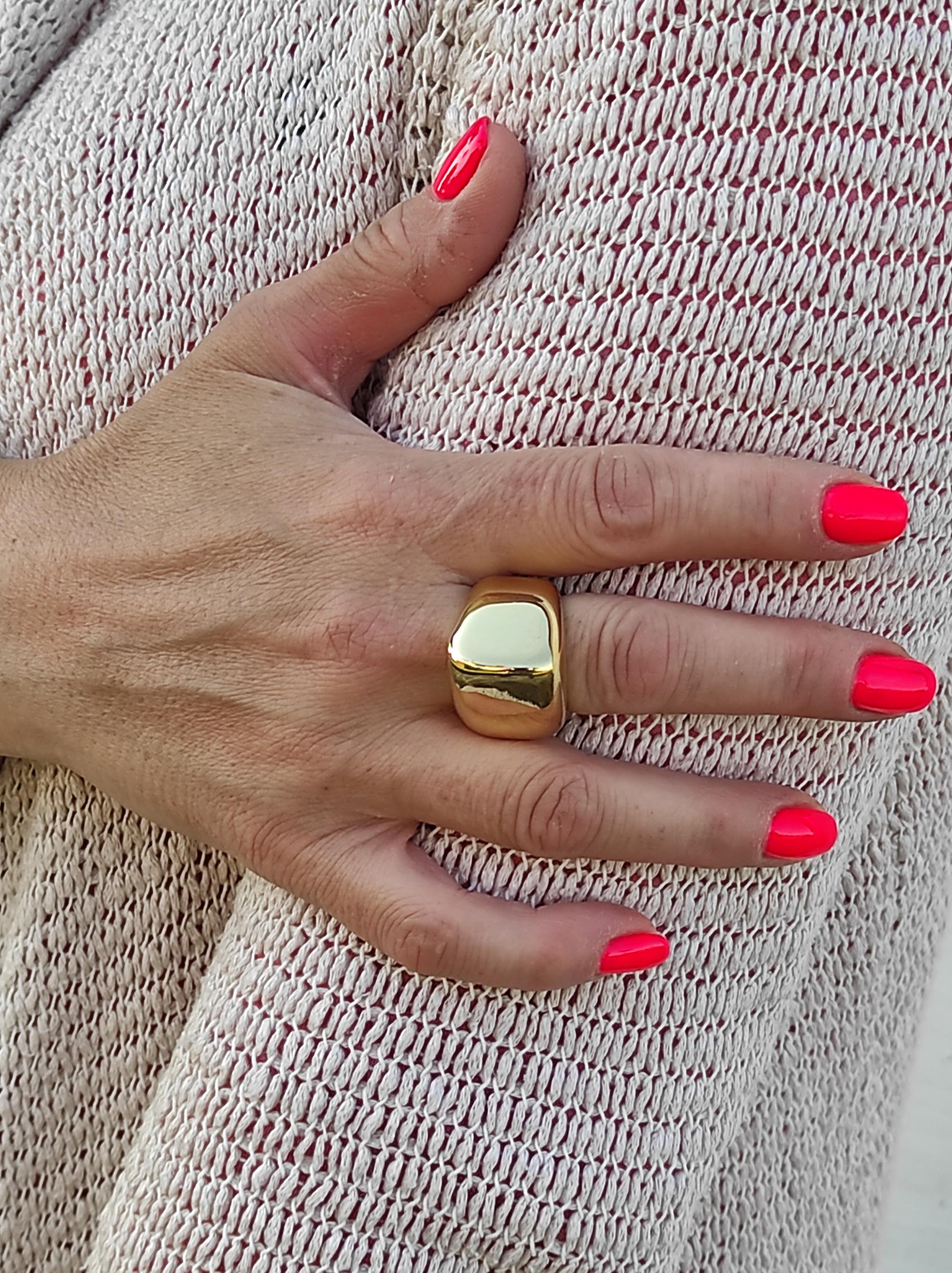 May include: A gold-colored ring with a rectangular shape is worn on a finger. The ring is smooth and shiny. The nails are painted a bright pink color. The background is a light-colored knitted fabric.