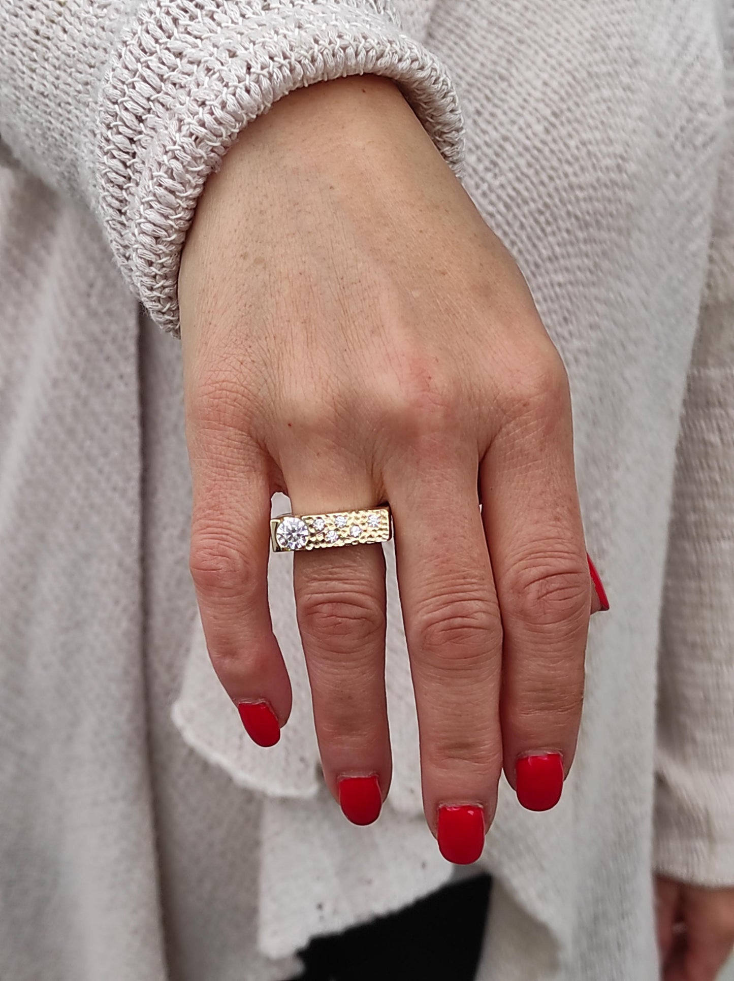 May include: A gold-toned ring with a rectangular band, featuring a large, clear gemstone and smaller accent stones. The ring is worn on a hand with red nail polish, set against a light-colored sweater.