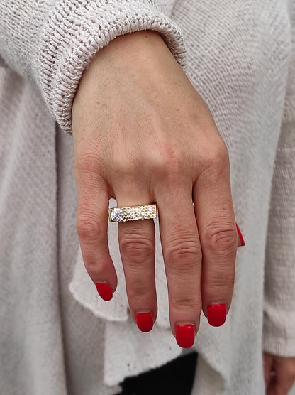 May include: A gold-toned ring with a rectangular band, featuring a large, clear gemstone and smaller accent stones. The ring is worn on a hand with red nail polish, set against a light-colored sweater.