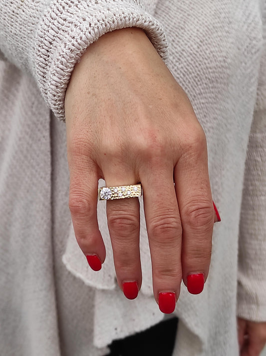 May include: A gold-toned ring with a rectangular band, featuring a large, clear gemstone and smaller accent stones. The ring is worn on a hand with red nail polish, set against a light-colored sweater.
