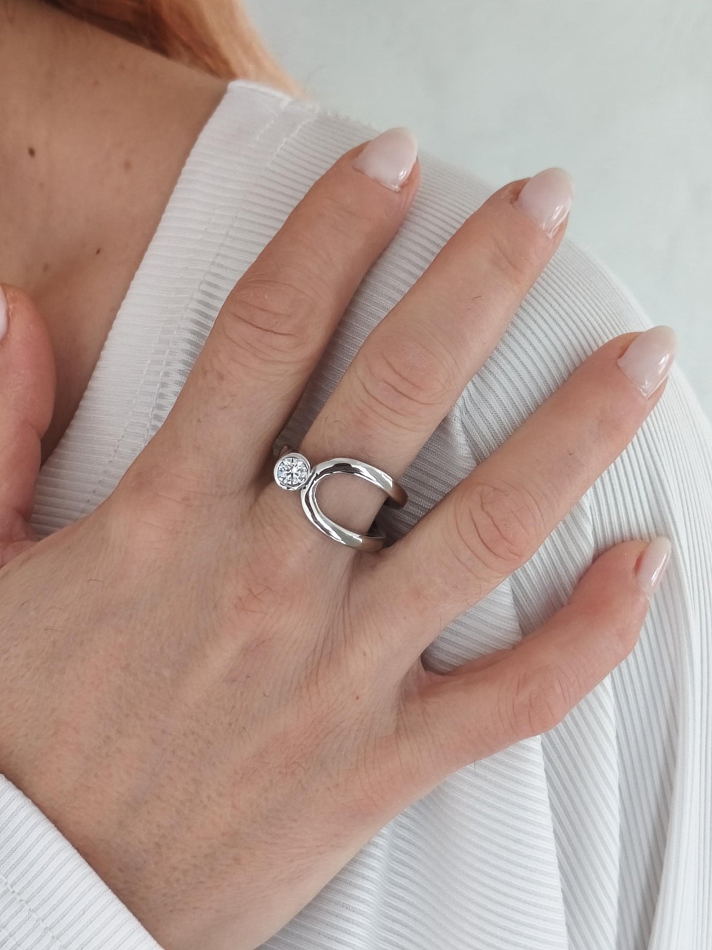 May include: A silver ring with a unique open design. The ring features a small, round, clear stone set on one side. The band curves around the finger, creating a modern and elegant look. The ring is worn on a hand.