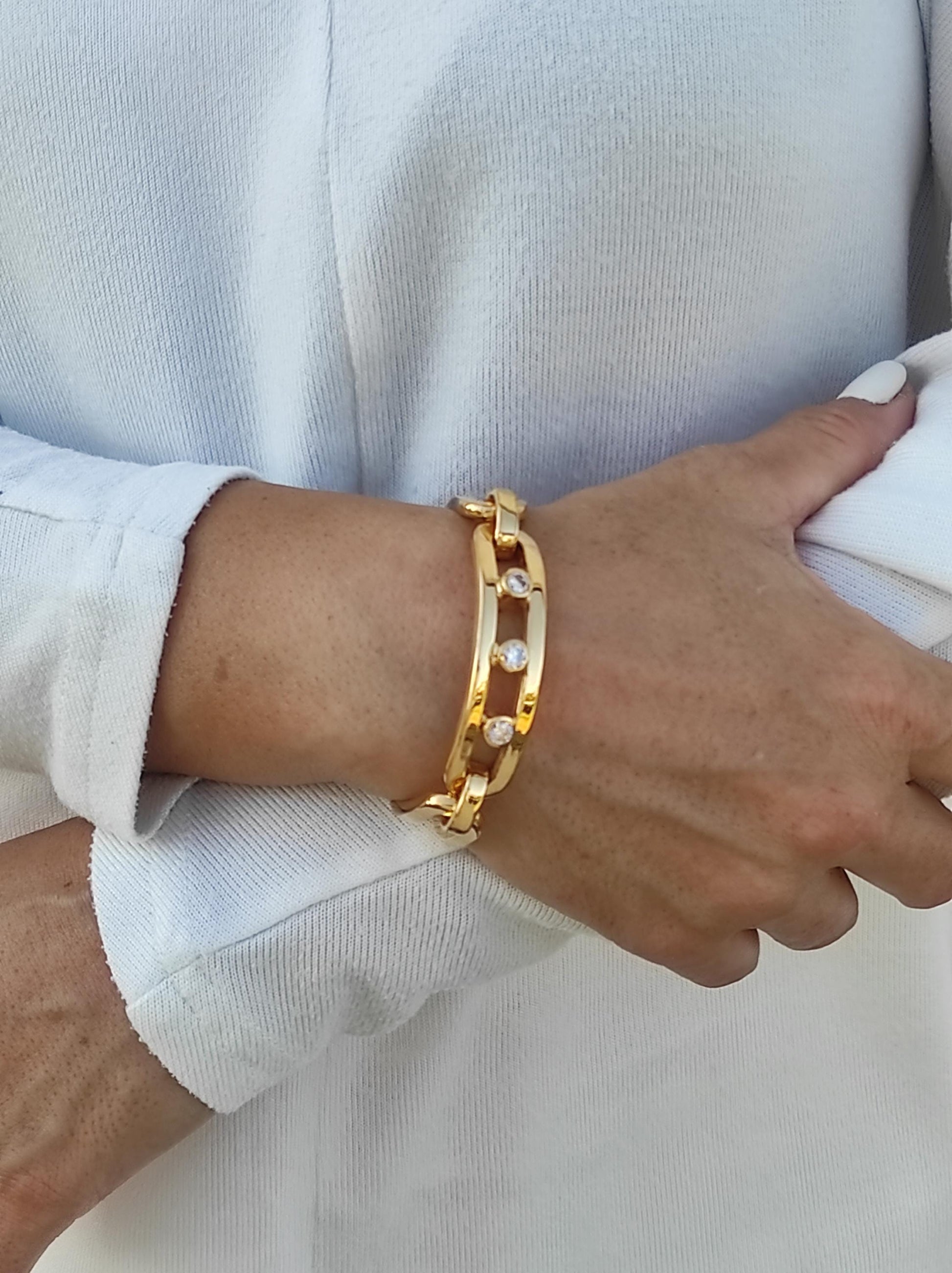 May include: A gold-colored bracelet with a chain link design. The bracelet features three clear, round gemstones set in the center of the links. The bracelet is worn on a wrist, with a white long-sleeved shirt in the background.
