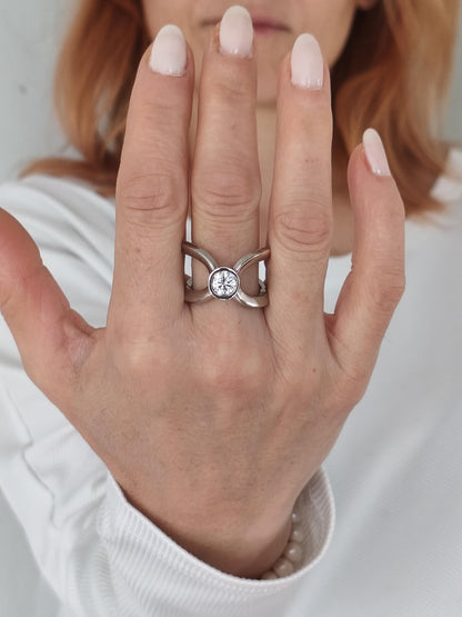 A silver ring with a round, clear gemstone at the center. The ring has a unique design with two bands crossing over each other. The ring is shown on a hand with light-colored nail polish.