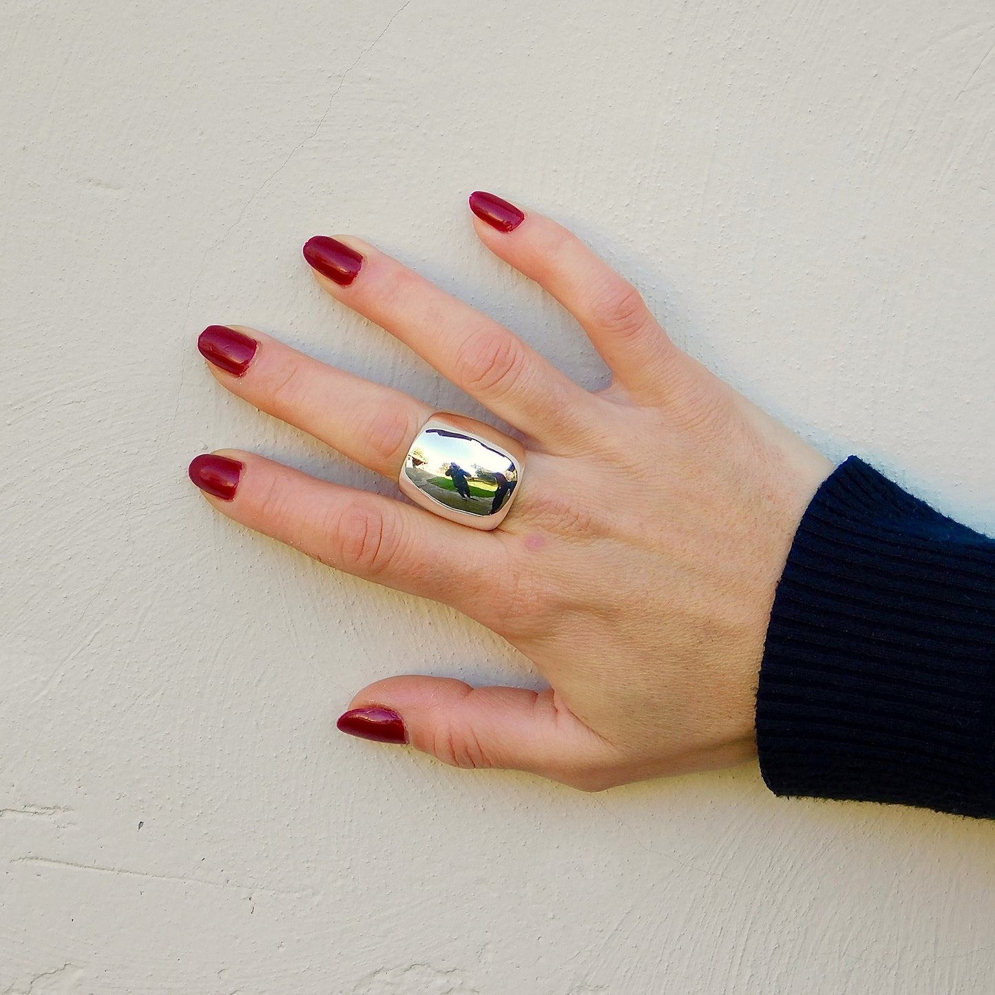 May include: A silver ring with a large, smooth, rectangular face. The ring is worn on the middle finger of a hand with red nail polish.