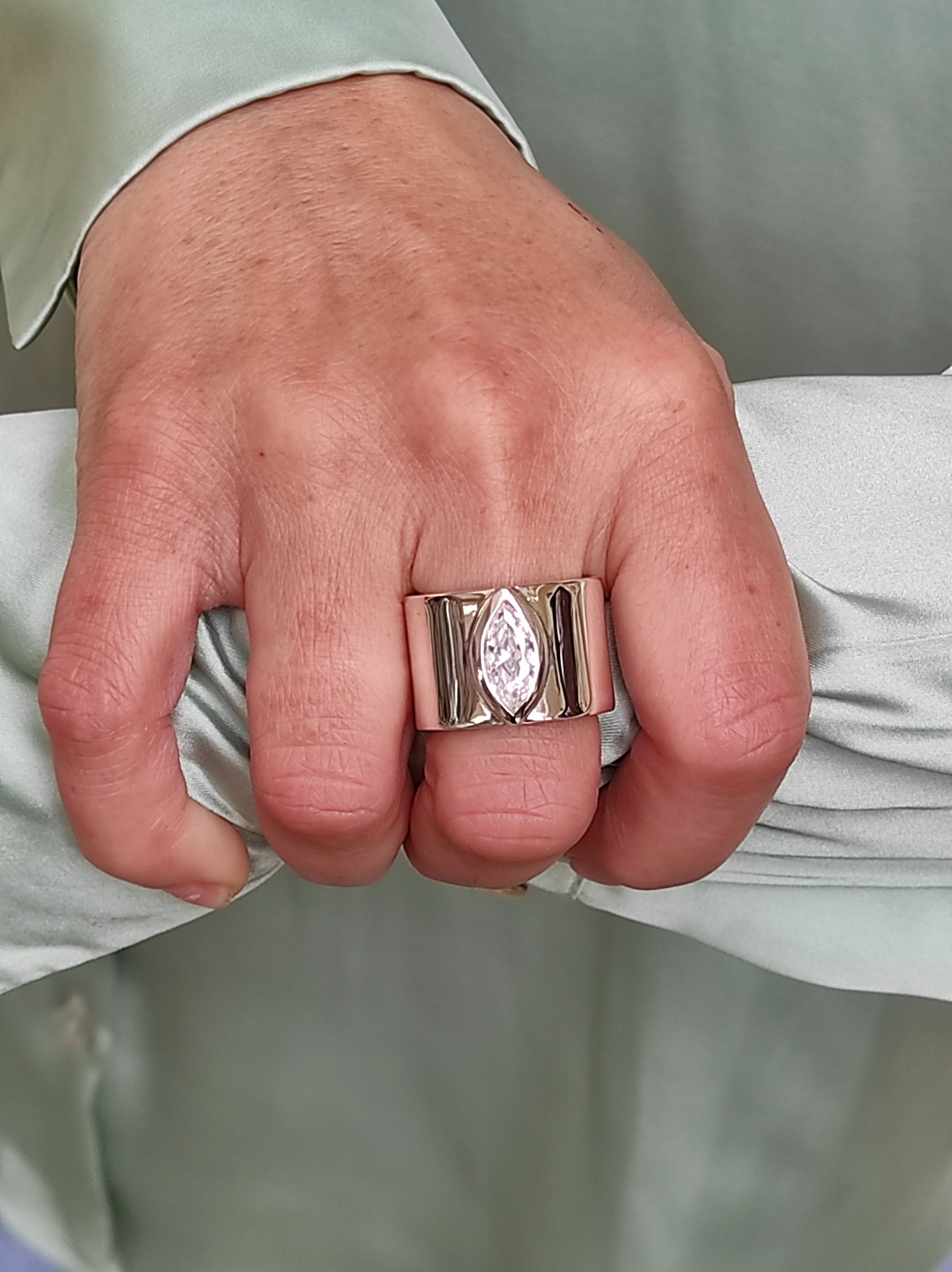 May include: A wide, gold-toned ring featuring a marquise-cut, clear gemstone. The ring has a thick band with vertical grooves on either side of the gemstone. The ring is worn on a finger, with a light green fabric in the background.