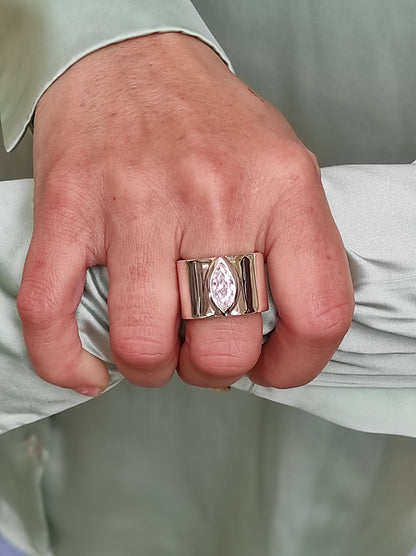 May include: A wide, gold-toned ring featuring a marquise-cut, clear gemstone. The ring has a thick band with vertical grooves on either side of the gemstone. The ring is worn on a finger, with a light green fabric in the background.