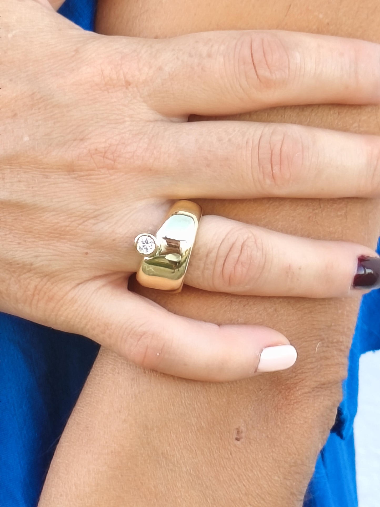 May include: A gold ring with a large, rounded band and a small, round diamond set on the side. The ring is worn on a finger, and the hand is visible against a blue background. The ring's design is simple and elegant.