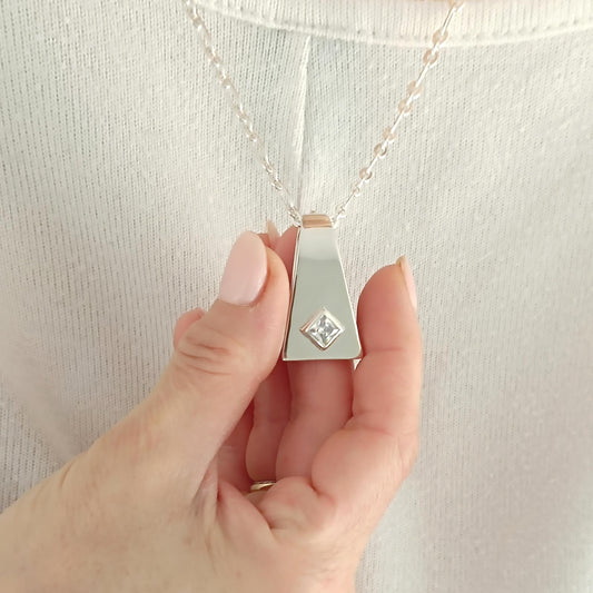 May include: Silver pendant necklace with a geometric design. The pendant has a trapezoid shape with a square, clear gemstone set in the center. The necklace is held by a person wearing a white shirt.