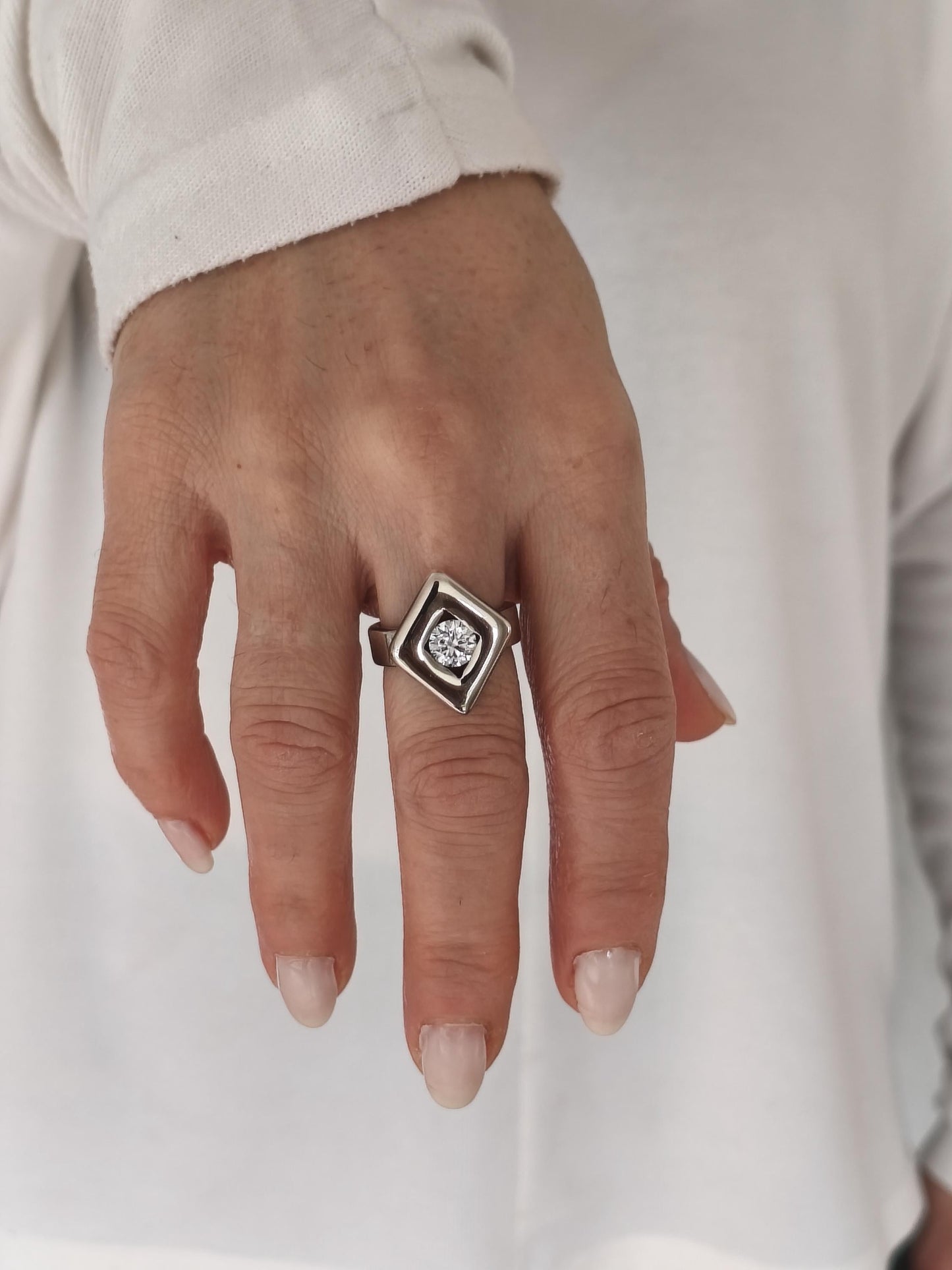 May include: A silver ring with a diamond-shaped setting. The ring features a central, round-cut diamond. The setting is a geometric design, with a square outer shape and a diamond-shaped inner frame. The ring is worn on a finger.