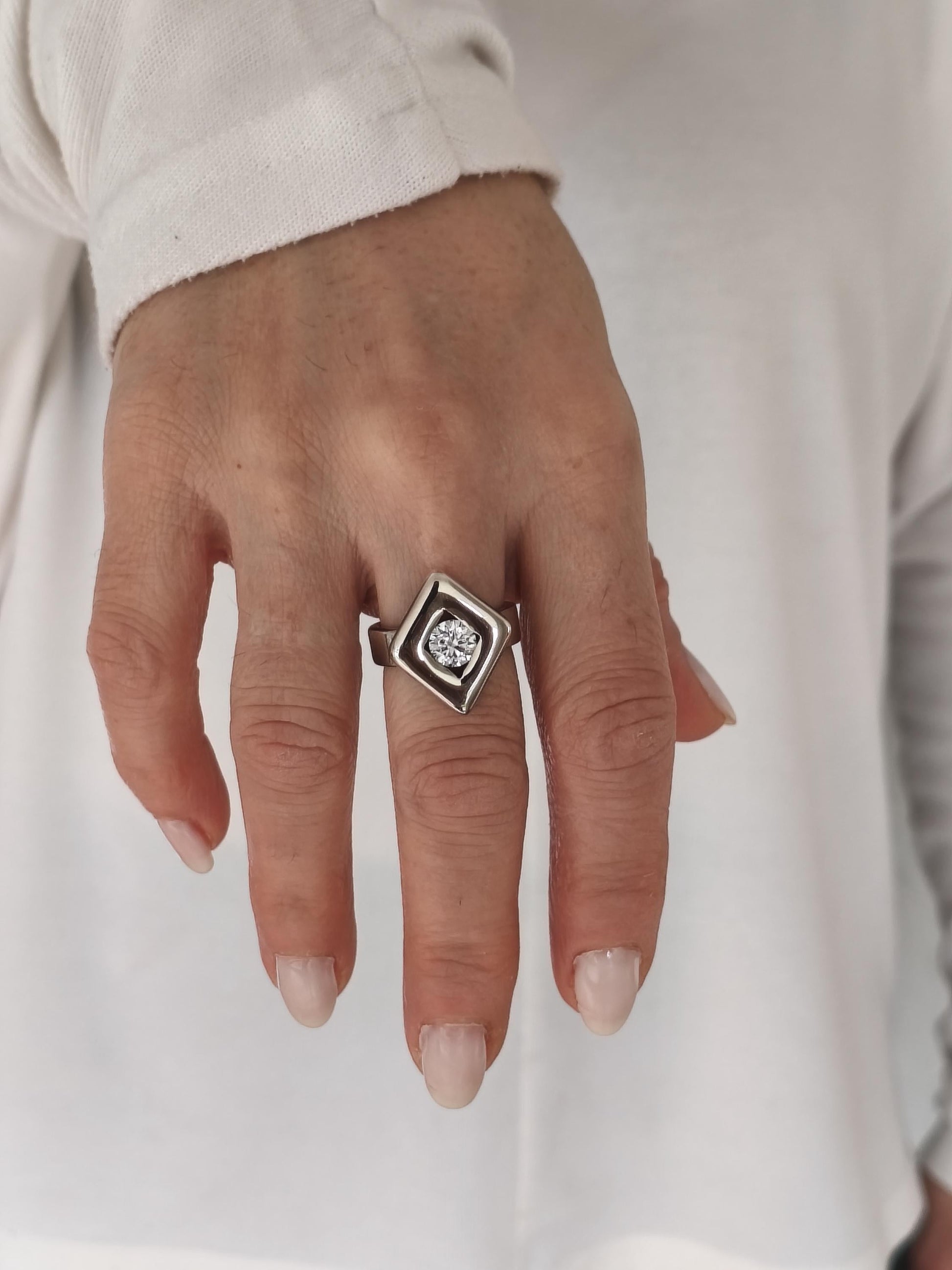 May include: A silver ring with a diamond-shaped setting. The ring features a central, round-cut diamond. The setting is a geometric design, with a square outer shape and a diamond-shaped inner frame. The ring is worn on a finger.