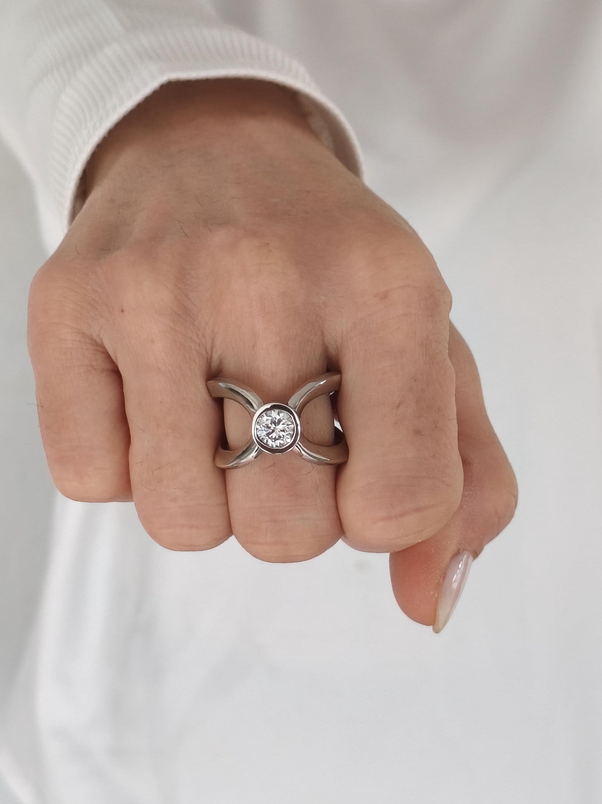 May include: A silver-toned ring with a large, round, clear gemstone at the center. The ring has a unique design with intersecting bands forming an X shape. The ring is shown on a hand.