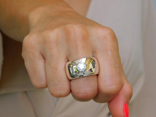 May include: A wide, polished silver ring with a large, clear gemstone set in the center. The ring has a rounded, rectangular shape and reflects light, creating a bright, eye-catching appearance. The gemstone is round and sparkles.