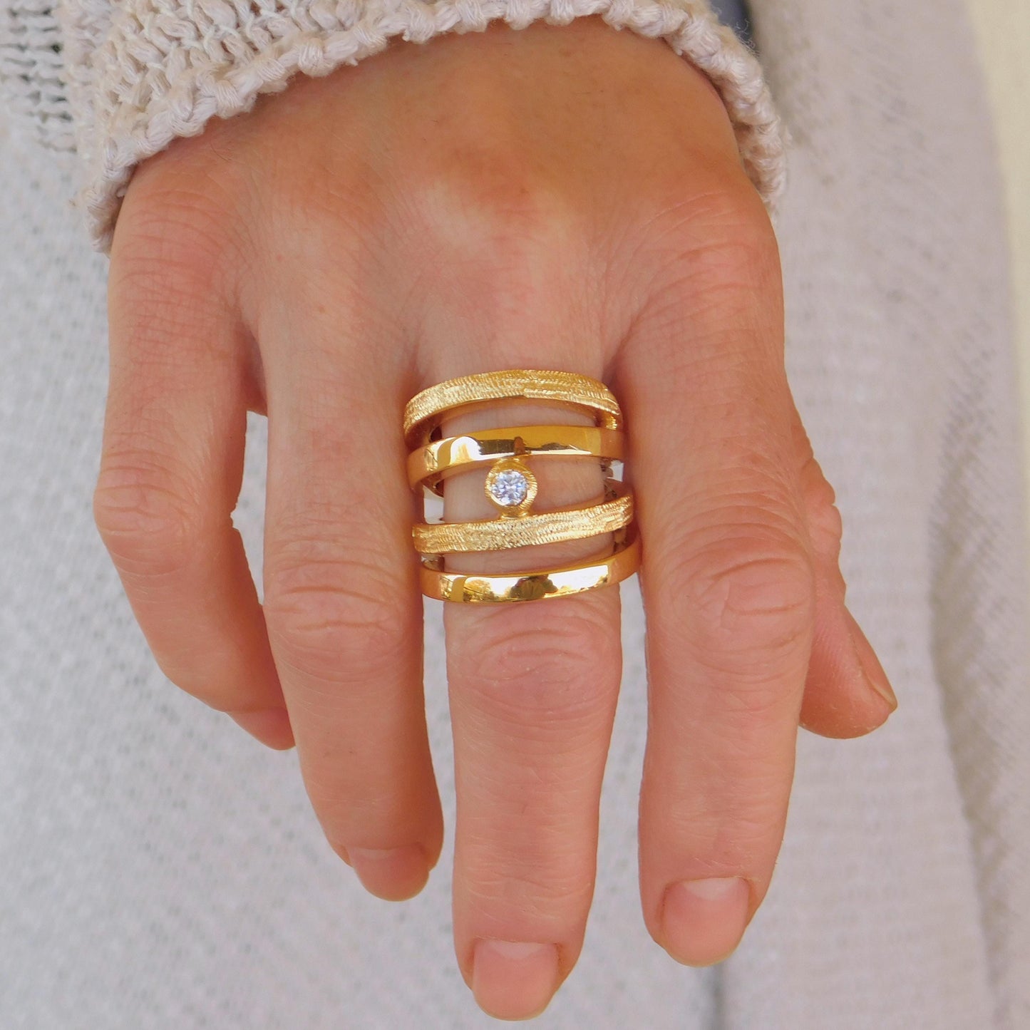 May include: A gold-colored ring with multiple bands, some textured, and a small, clear gemstone. The ring is worn on a finger, showcasing its design and craftsmanship. The bands have a textured surface.