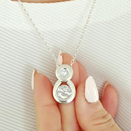 May include: A silver necklace with a pendant featuring two clear, round gemstones. The pendant is suspended from a delicate chain. The gemstones are set in a silver bezel setting. The necklace is displayed against a white background.