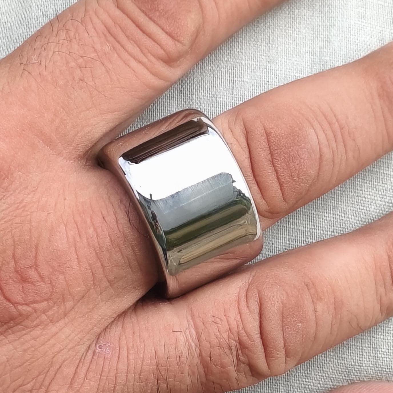 May include: A wide, polished silver ring with a rectangular design. The ring is worn on a finger, reflecting light. The jewelry piece has a smooth, shiny surface. This ring is a bold statement piece.