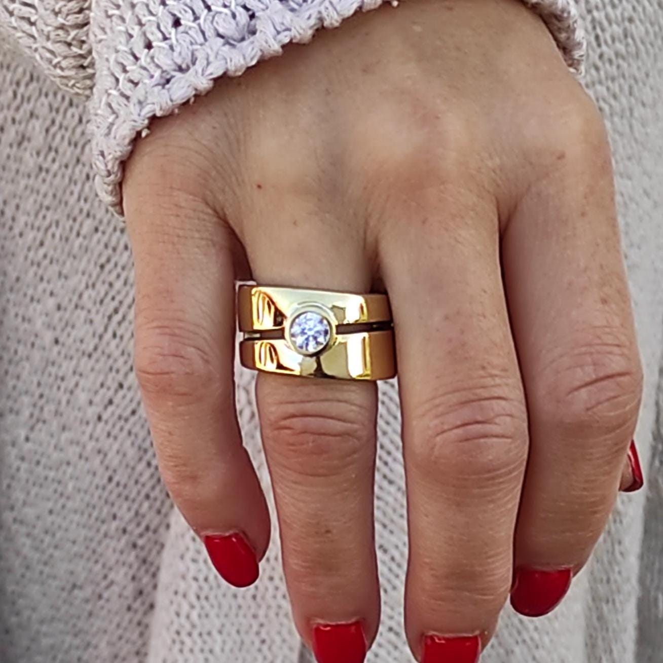 May include: A gold ring with a single, round, clear stone set in the center. The ring is made of two bands, one wider than the other, and is worn on the ring finger of a hand.