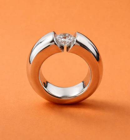 May include: A polished silver ring with a large, clear diamond set in the center. The ring has a thick band and a modern, minimalist design. The background is a solid orange color.