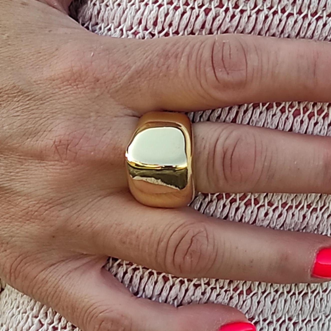 May include: A gold-colored ring with a rectangular shape is worn on a finger. The ring is smooth and shiny. The nails are painted a bright pink color. The background is a light-colored knitted fabric.