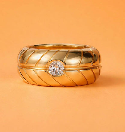 May include: A gold-colored ring with a central, clear gemstone. The ring has a thick band with a textured, diagonal pattern. The background is a solid orange color, providing a contrast to the ring's metallic sheen.