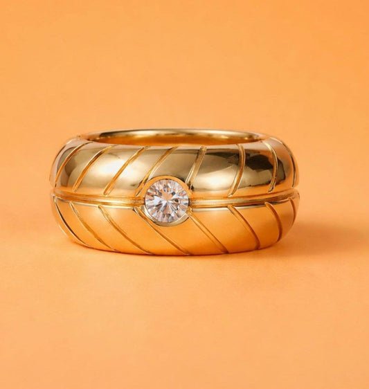 May include: A gold-colored ring with a central, clear gemstone. The ring has a thick band with a textured, diagonal pattern. The background is a solid orange color, providing a contrast to the ring's metallic sheen.