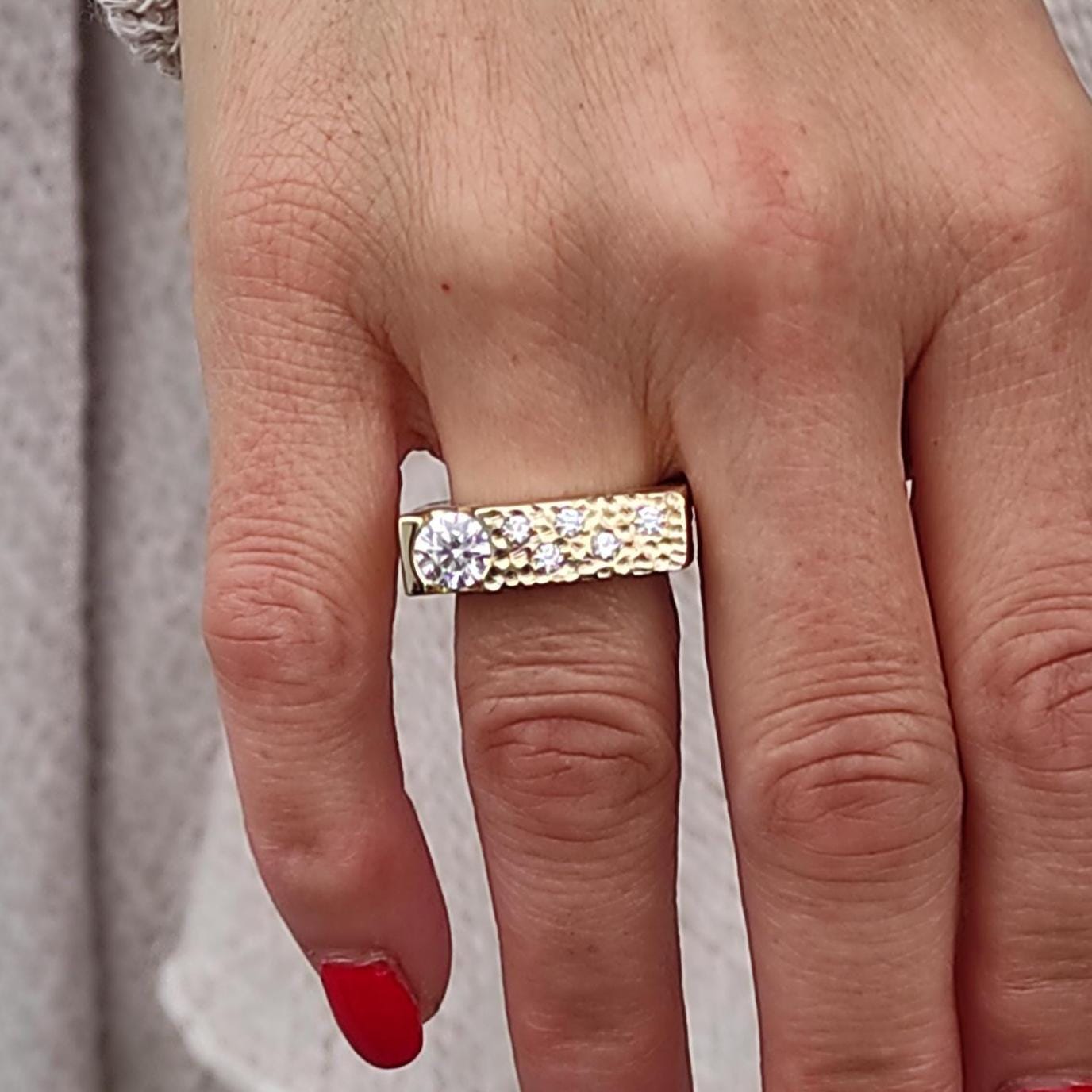 May include: A gold-toned ring with a rectangular band, featuring a large, clear gemstone and smaller accent stones. The ring is worn on a hand with red nail polish, set against a light-colored sweater.