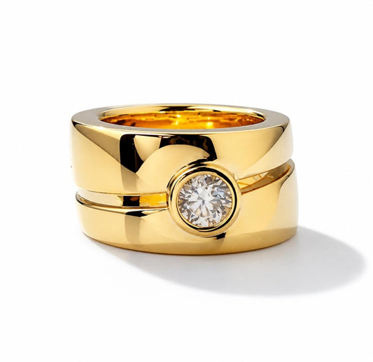 May include: A wide, polished yellow gold band ring featuring a split design with a single, round, brilliant-cut clear gemstone set in a bezel setting at the center. The ring has a smooth, reflective surface and a thick, substantial profile.