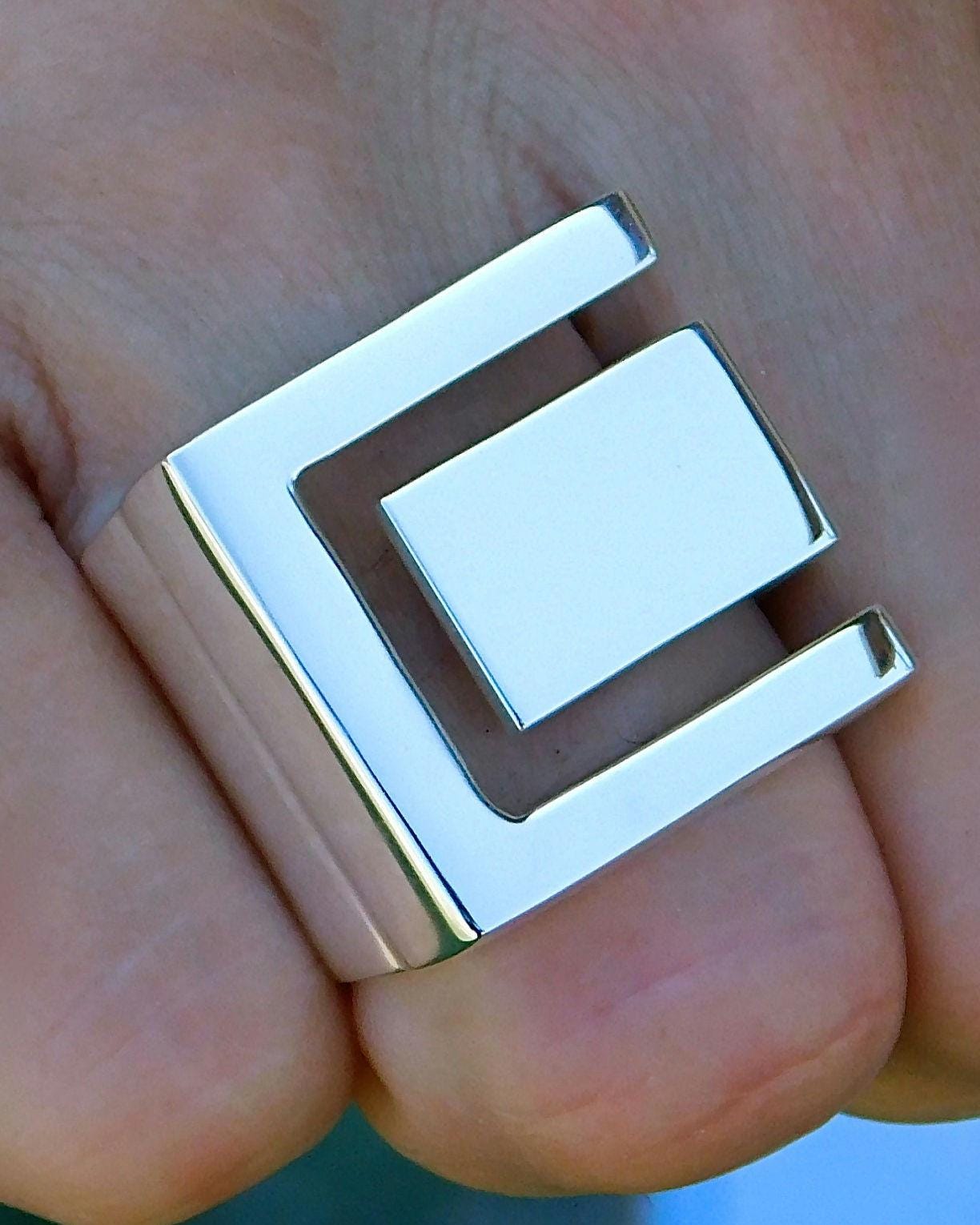 May include: A silver ring with a square design featuring a cutout in the center. The ring is worn on a finger.