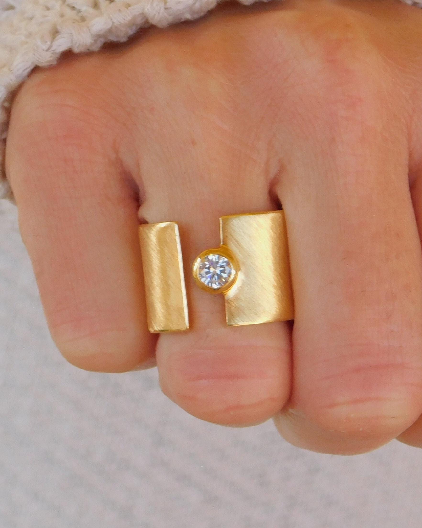 May include: A gold ring with a single, clear gemstone set in the center. The ring is made of two rectangular pieces of metal that are joined together.